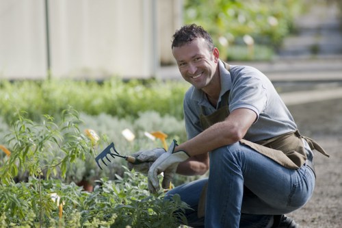 The image depicts a smiling man in casual clothing and gloves crouching in a garden. He is tending to a bed of green plants and flowers, which include some yellow and orange blooms. The garden features a lawn area with dense, well-maintained grass in the foreground, bordered by a paved pathway. In the background, there are a variety of shrubs and small trees, with sunlight filtering through the leaves, indicating a clear day. The setting appears to be a pleasant outdoor space, possibly a backyard or community garden, with a natural and tidy layout. The man is using a small gardening fork, suggesting he is engaged in planting or weeding activities. This scene exemplifies routine garden maintenance, aligning with services offered by companies like Garden Maintenance Bayswater, and highlights typical elements found in landscaped outdoor areas within the Bayswater or nearby London region.