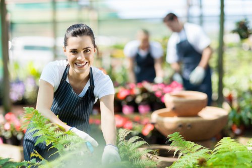 A smiling young woman wearing a blue and white striped apron and gardening gloves is working in a bright, spacious greenhouse or garden centre. She is kneeling or bending over a bed of lush, green ferns and small plants, which are in the foreground of the image. Behind her, two other gardeners or staff members, dressed similarly in aprons and gloves, are tending to colourful potted flowers, possibly pink or red blooms, arranged on garden tables or display shelves. The background features a well-lit environment with transparent large panels allowing natural light to illuminate the space. The environment appears warm and welcoming, suitable for outdoor or greenhouse gardening activities. The setting reflects professional gardening work, likely associated with landscaping or plant care services provided by Garden Maintenance Bayswater, supporting local gardening and landscaping needs in Bayswater or nearby areas in London, under the postcode W2. The scene demonstrates tools and activities typical of garden maintenance, set in a professionally maintained and visually appealing outdoor and indoor plant environment.
