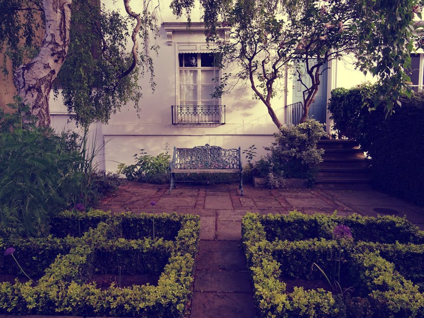 A small, well-maintained Bayswater garden featuring a lush green lawn with neatly trimmed grass, bordered by flower beds containing colorful flowering plants and leafy shrubs. In the background, a tal